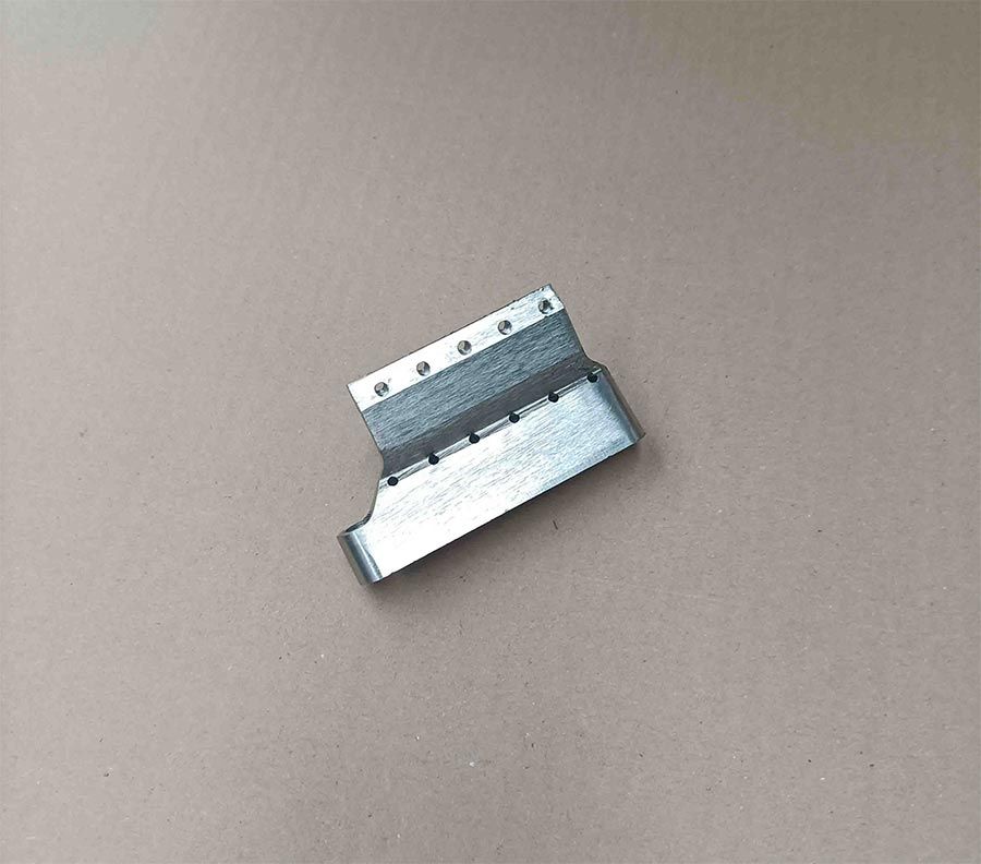 Soporte metálico con múltiples agujeros, plegado en forma de L, apoyado sobre una superficie de color beige.