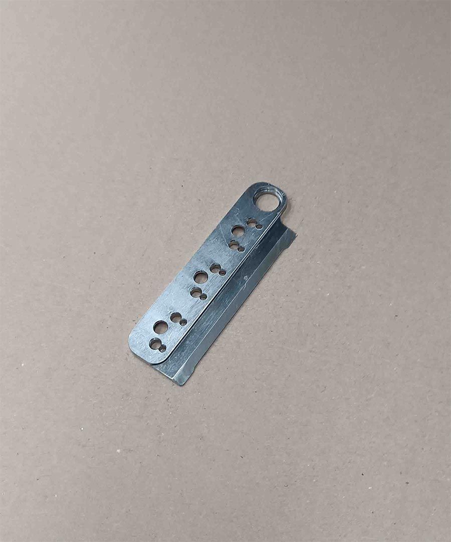 Una pequeña hoja de metal plateada con múltiples agujeros y un lazo en el extremo, colocada sobre una superficie de color beige.