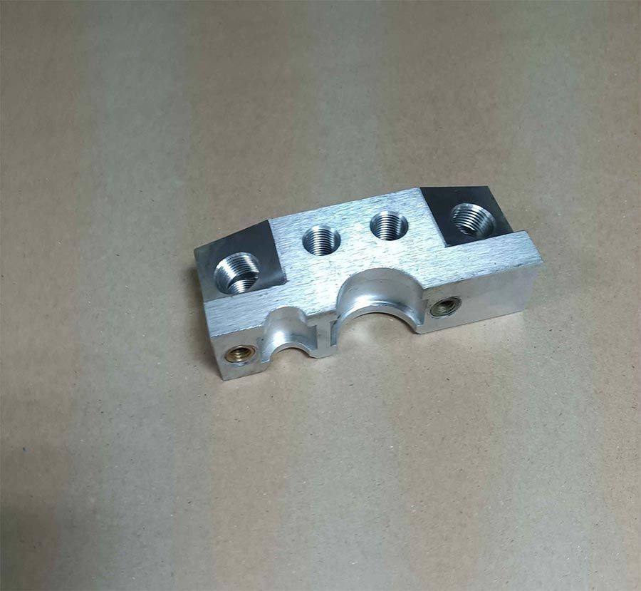 Bloque de metal plateado con múltiples orificios, una hendidura curva y puntos de montaje.