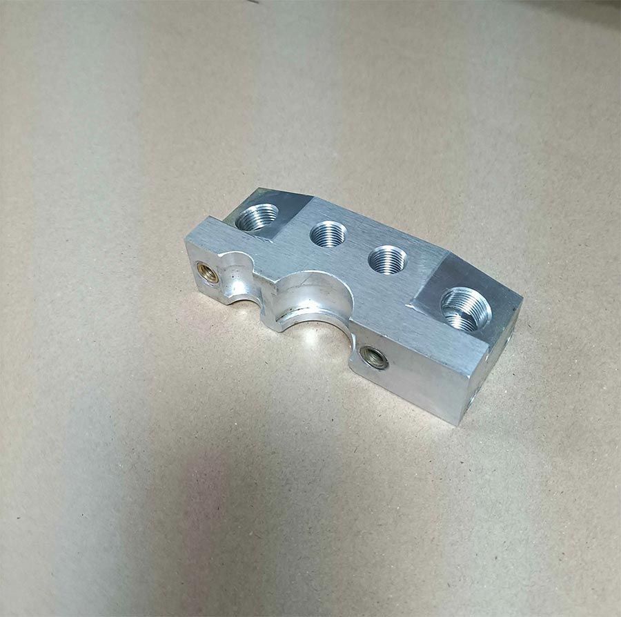 Pieza mecanizada en metal plateado con agujeros y tramos curvados, sobre superficie neutra.