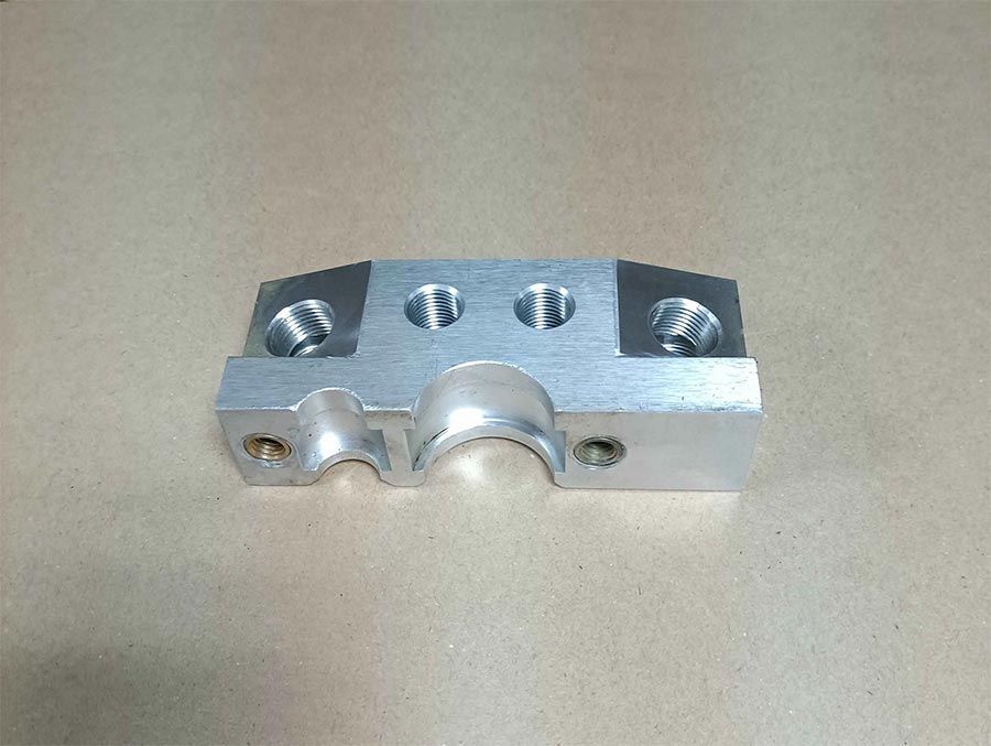 Bloque de metal de color plateado con agujeros y secciones curvas, probablemente un componente mecánico.