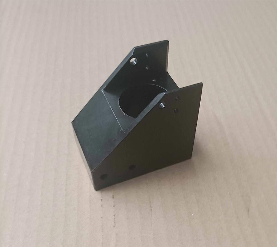 Soporte metálico negro, de forma angular y con abertura circular. Se fija con tornillos pequeños y se coloca sobre una superficie marrón.