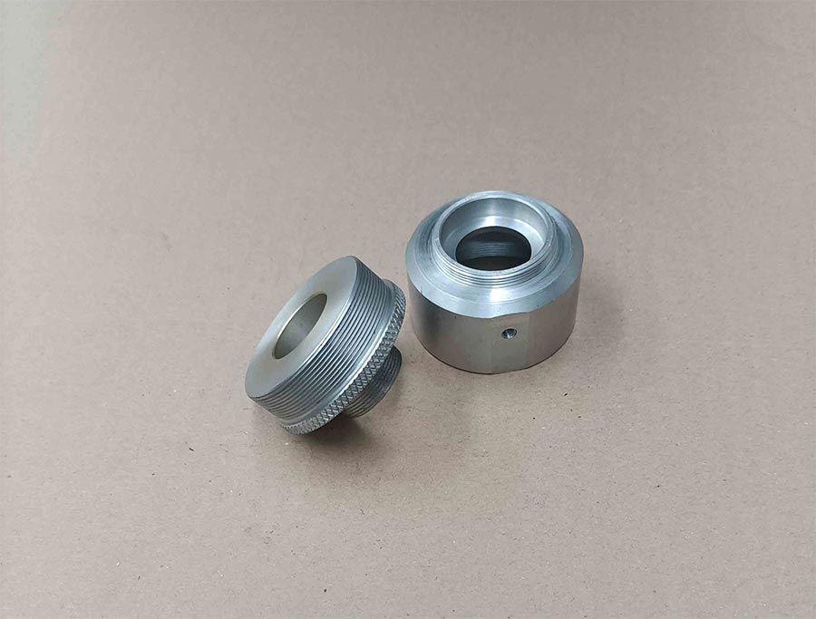 Dos piezas metálicas mecanizadas, una de ellas con borde moleteado, sobre una superficie beige.