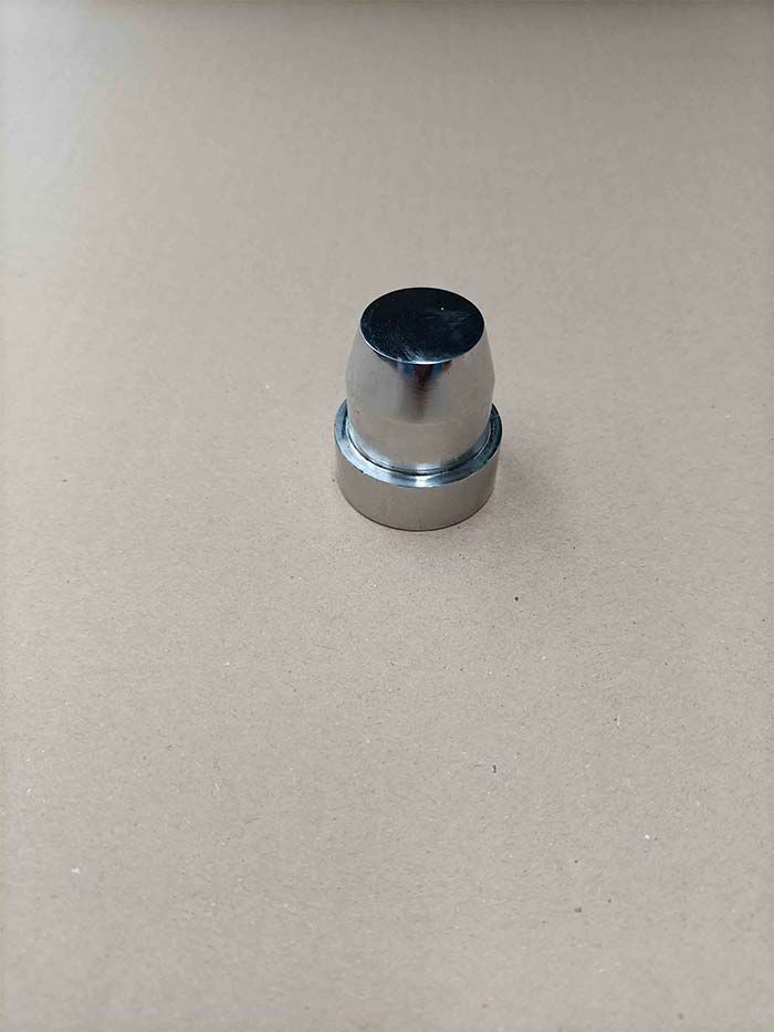 Objeto metálico brillante, cilíndrico, con la parte superior ensanchada y una base circular, sobre una superficie de color canela.