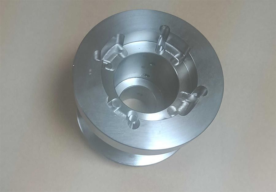 Pieza de metal mecanizada en plata con orificio central y recortes circundantes.