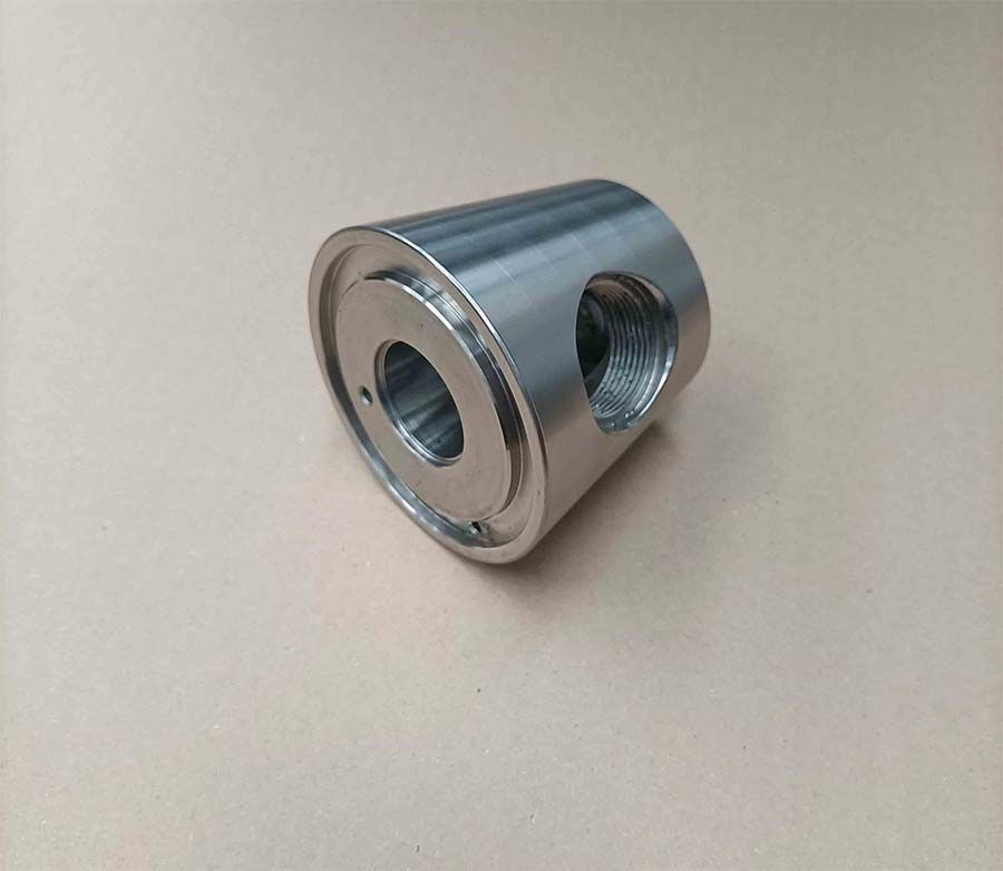 Pieza cilíndrica de metal plateado con dos orificios laterales y un orificio central sobre una superficie de color claro.