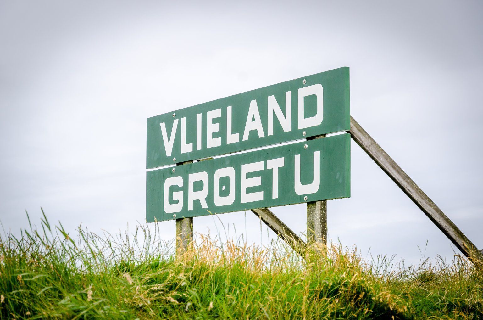 Een groen bord met de tekst vlieland groet u staat op de top van een met gras begroeide heuvel.