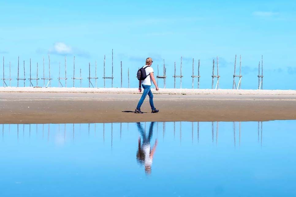 Een man loopt met een rugzak over het strand en zijn weerspiegeling is in het water.