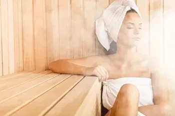 Een in een handdoek gewikkelde vrouw zit in een houten sauna met haar ogen dicht.