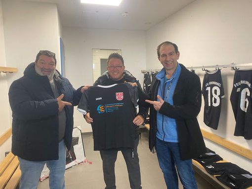 Drei Männer in einer Umkleidekabine, zwei zeigen auf ein Trikot, das der mittlere Mann hochhält. Schwarzes Trikot mit weißem Saum und Logo.