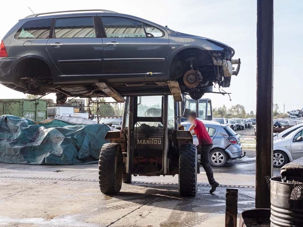 Voiture soulevée par un chariot élévateur dans un parc automobile, un ouvrier se tenant à côté.