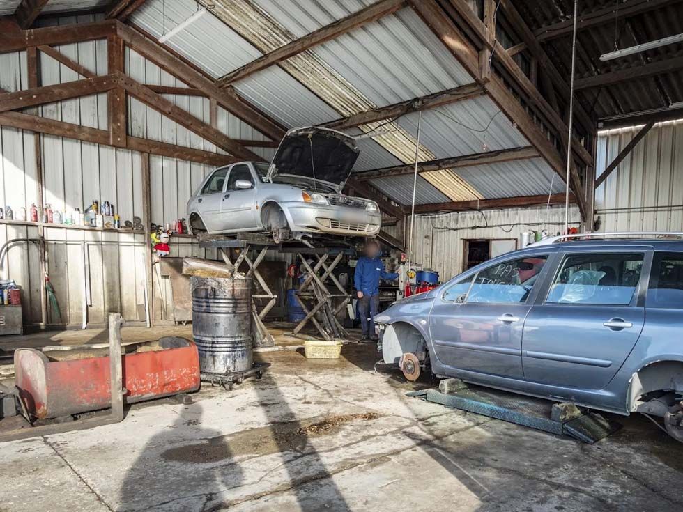 Voitures sur des ponts élévateurs dans un garage automobile encombré, avec une voiture bleue au premier plan et une voiture argentée surélevée à l'arrière-plan.