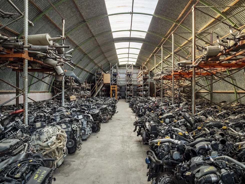 Allée d'entrepôt remplie de moteurs et de pièces de moto empilés sous un toit industriel incurvé