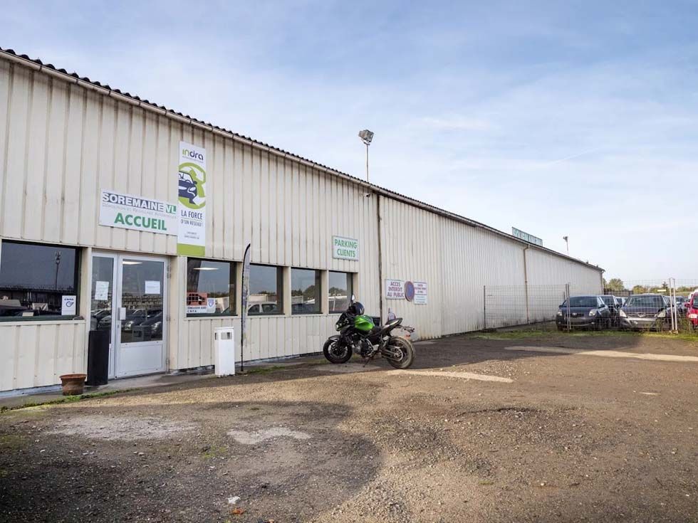 Long bâtiment industriel blanc avec une moto garée à l'extérieur sur un terrain en gravier