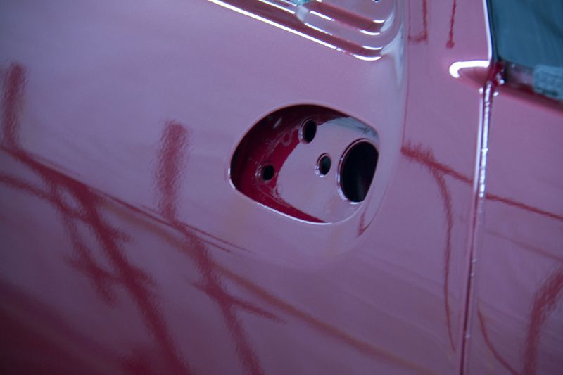 Gros plan sur une portière de voiture rose dont la poignée a été retirée, laissant apparaître l'emplacement de fixation.