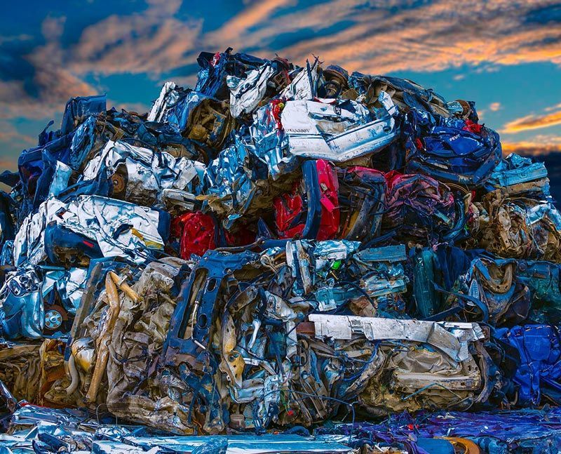 Des voitures broyées empilées dans une casse, avec des débris de métal bleu, rouge, blanc et or sous un ciel bleu.