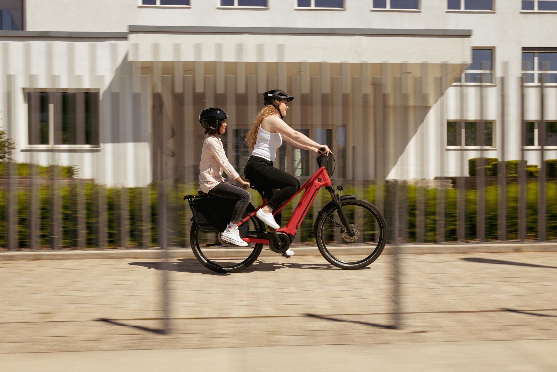 Zwei Personen mit Helmen fahren mit einem roten E-Bike auf einem Weg vor einem modernen weißen Gebäude entlang.