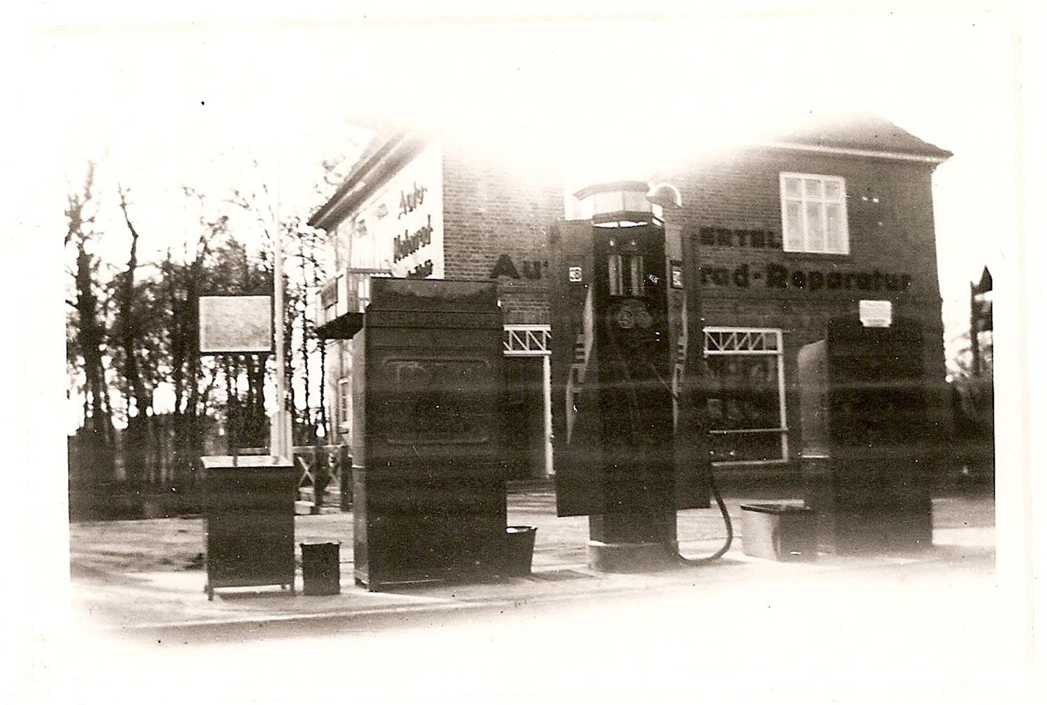 Ein historisches, sepiafarbenes Foto einer kleinen Tankstelle am Straßenrand mit drei Zapfsäulen vor einem Backsteingebäude.