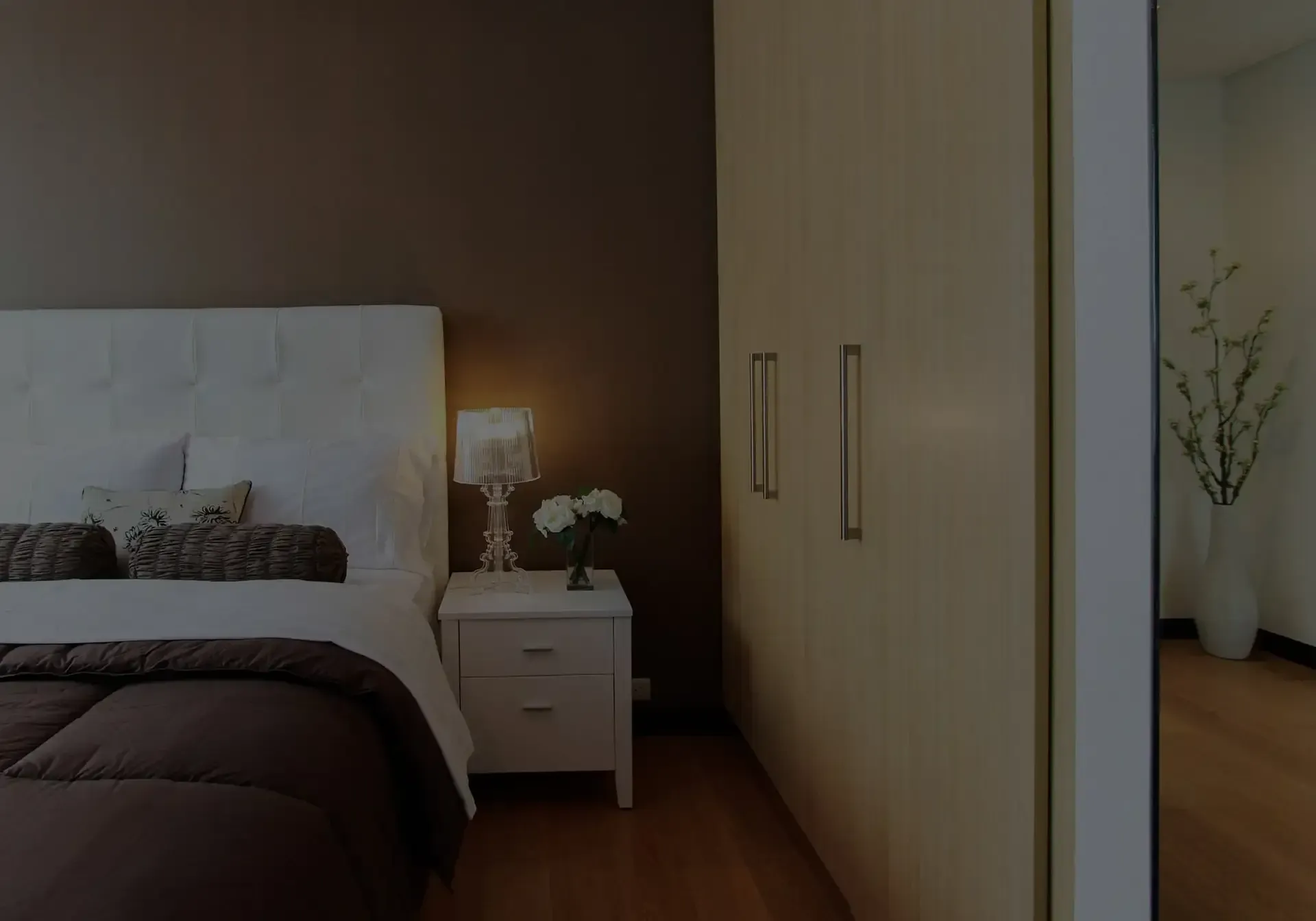 Modernes Schlafzimmer mit weissem, gestepptem Bett, Nachttischlampe und verspiegeltem Kleiderschrank in warmen Holztönen