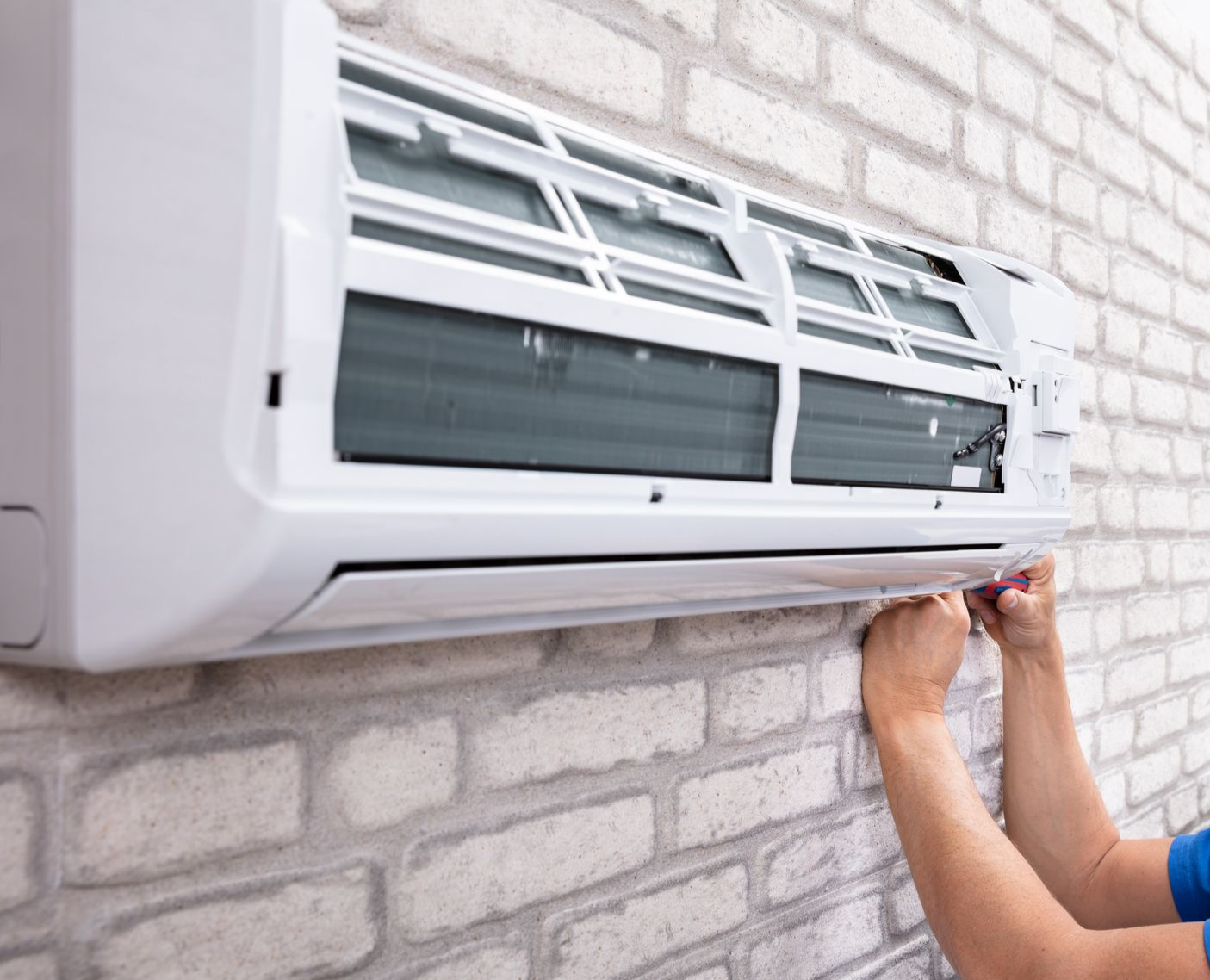 Les mains d'un ouvrier ajustent le couvercle d'un climatiseur mural blanc contre un mur de briques blanches.