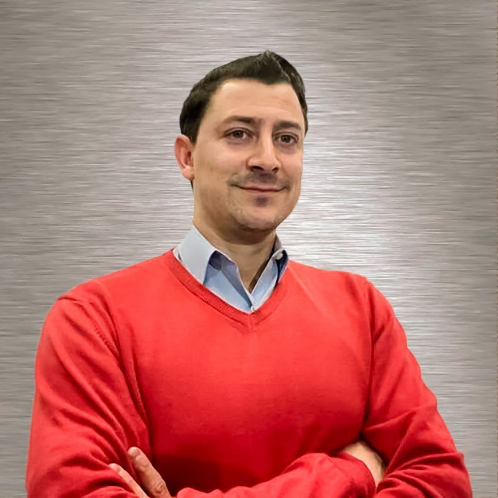 Hombre con los brazos cruzados, vestido con suéter rojo y camisa de cuello azul claro, fondo neutro.