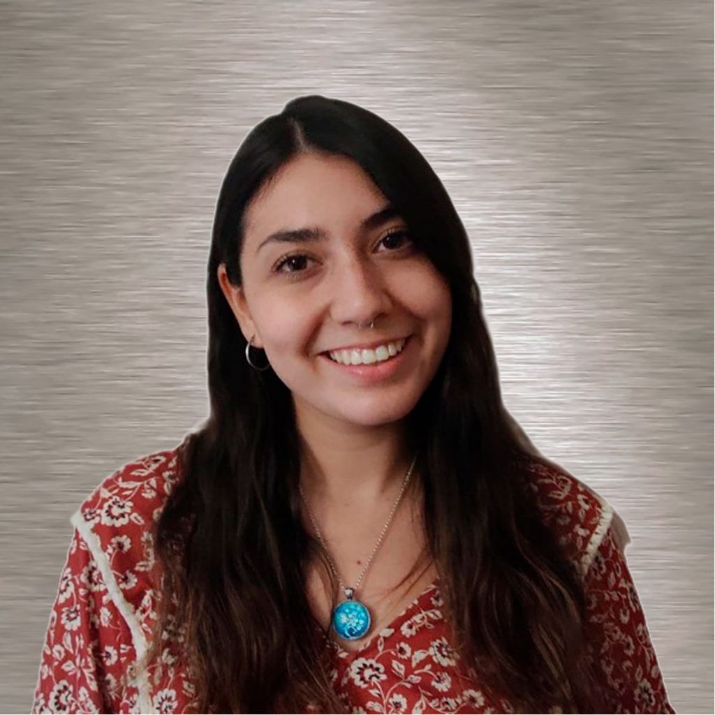 Una mujer de cabello oscuro sonríe, vistiendo un top y un collar floral, frente a un fondo gris.