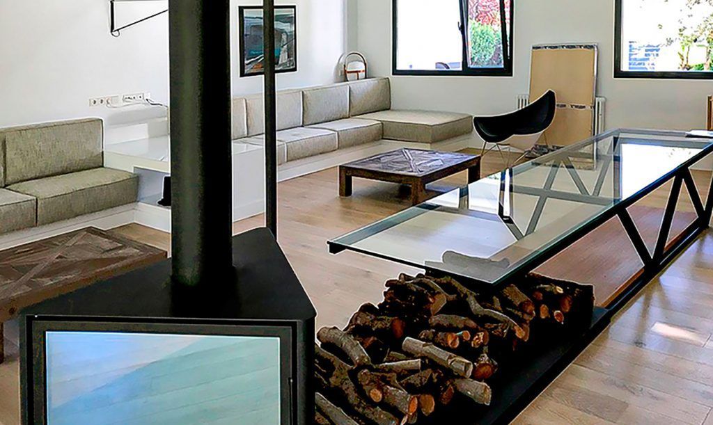 Sala de estar con chimenea moderna, leñero incorporado, mesa con tapa de vidrio, sofás seccionales beige y ventanas.