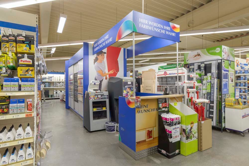 Gang im Baumarkt, Auslage mit Farbe in den Farben Blau und Weiß. Person malt auf einer Tafel.