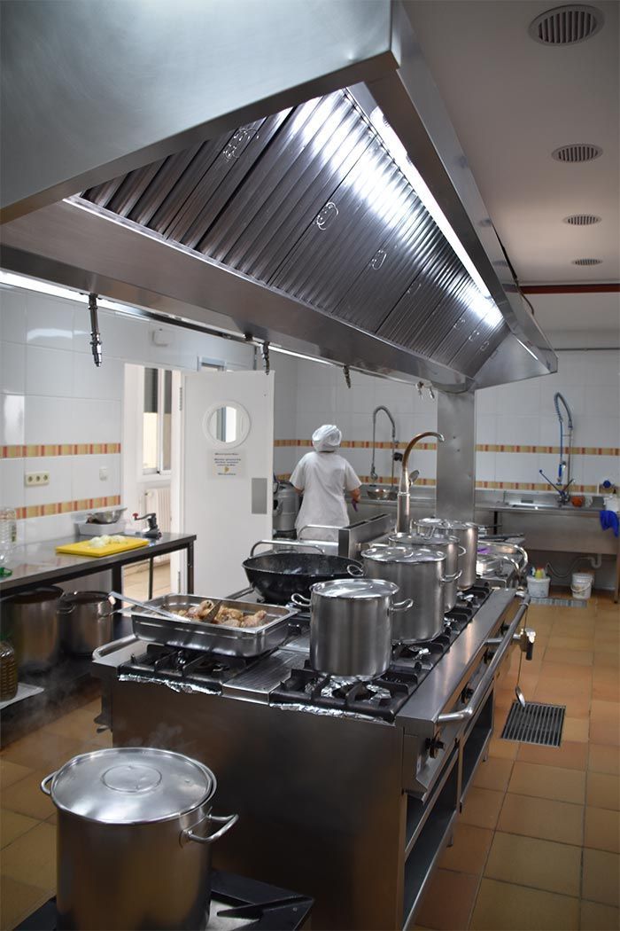 Una cocina de acero inoxidable con una gran campana extractora sobre una cocina industrial, ollas en la estufa y una persona trabajando en la parte de atrás.