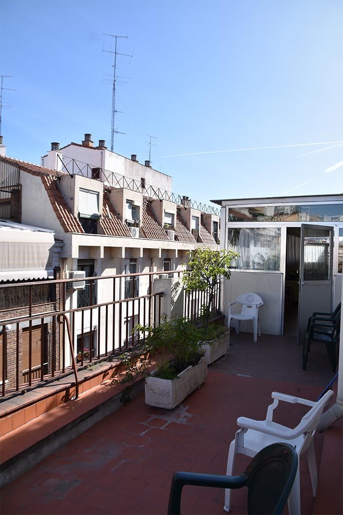 Una soleada terraza en la azotea, en un entorno urbano, con sillas de plástico blancas, plantas en macetas y edificios circundantes.