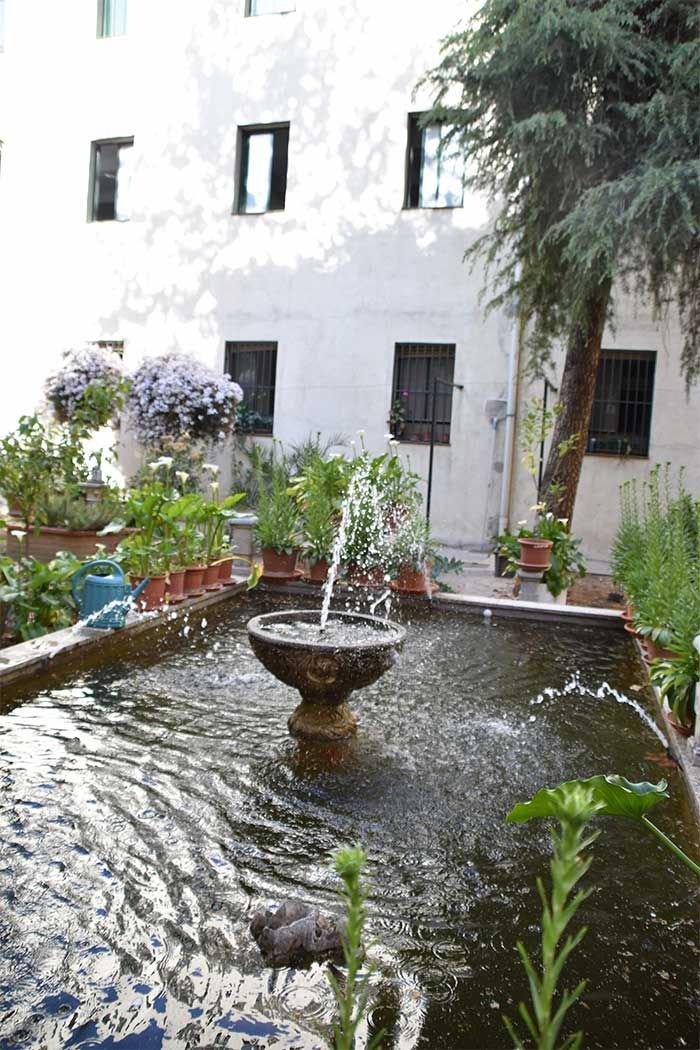 Una fuente de piedra vierte agua en una piscina rectangular situada en un patio frente a un edificio blanco con varias ventanas.