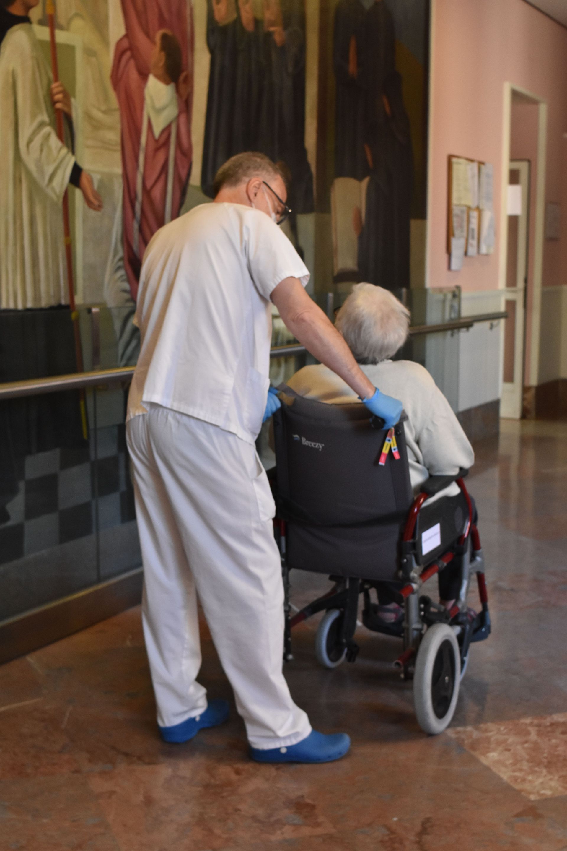 Un trabajador sanitario, vestido con uniforme quirúrgico y guantes azules, empuja a una persona en silla de ruedas a través de un pasillo decorado con murales.