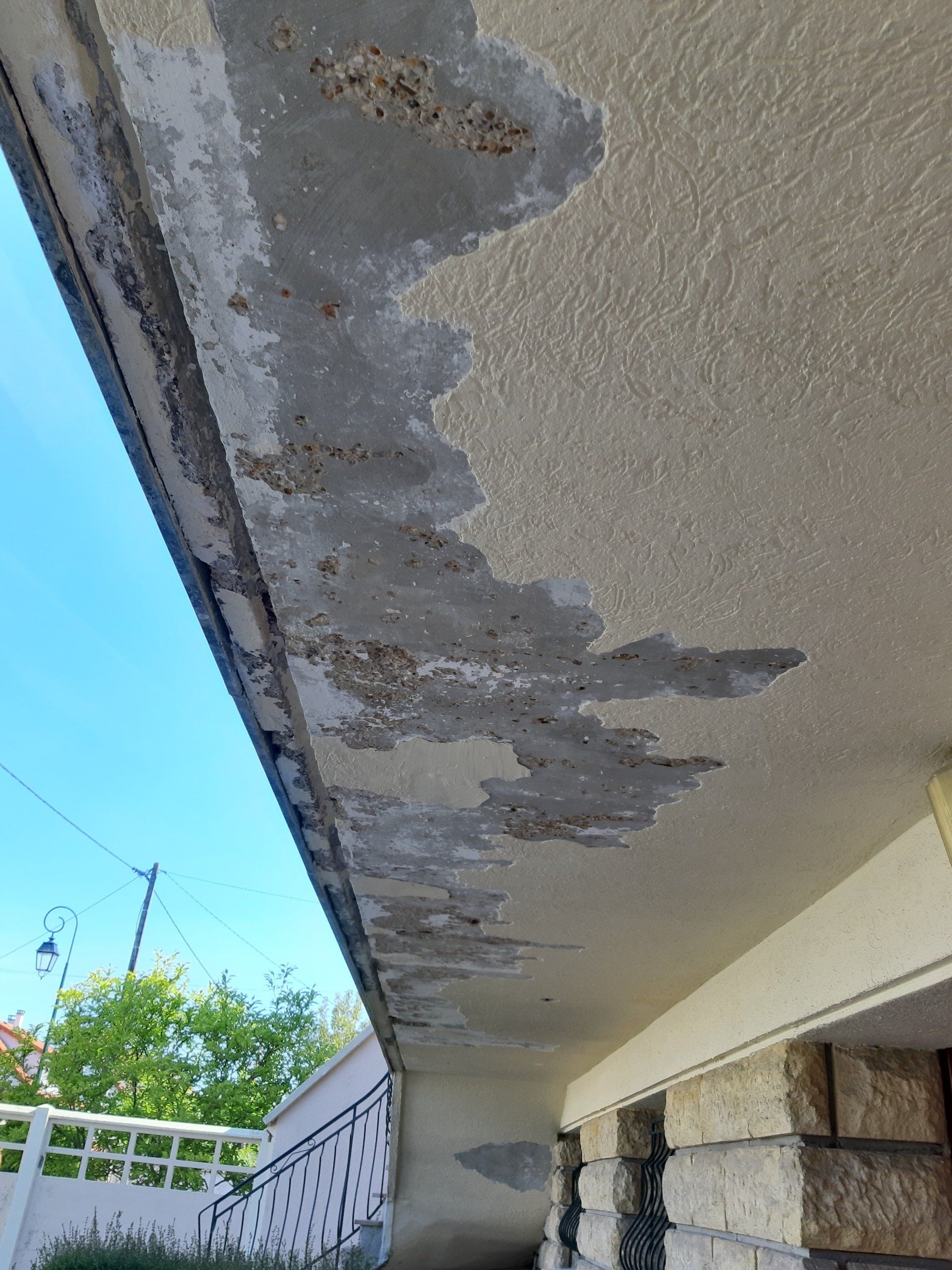 Plafond en béton endommagé, peinture écaillée, vu du dessous. Ciel bleu visible au-dessus.