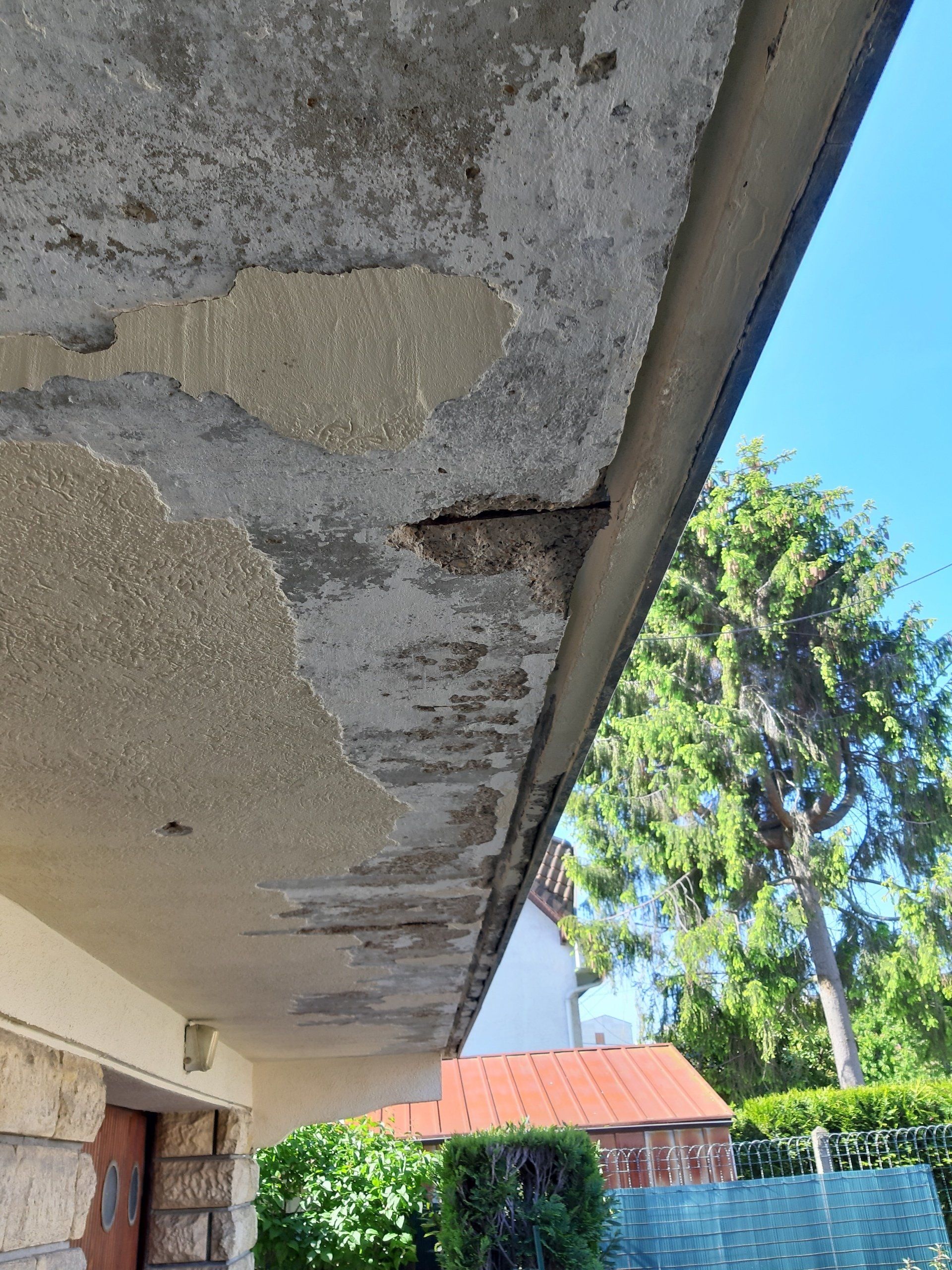 Auvent en béton endommagé avec des sections qui s'effondrent; vue extérieure avec un ciel bleu et des arbres.