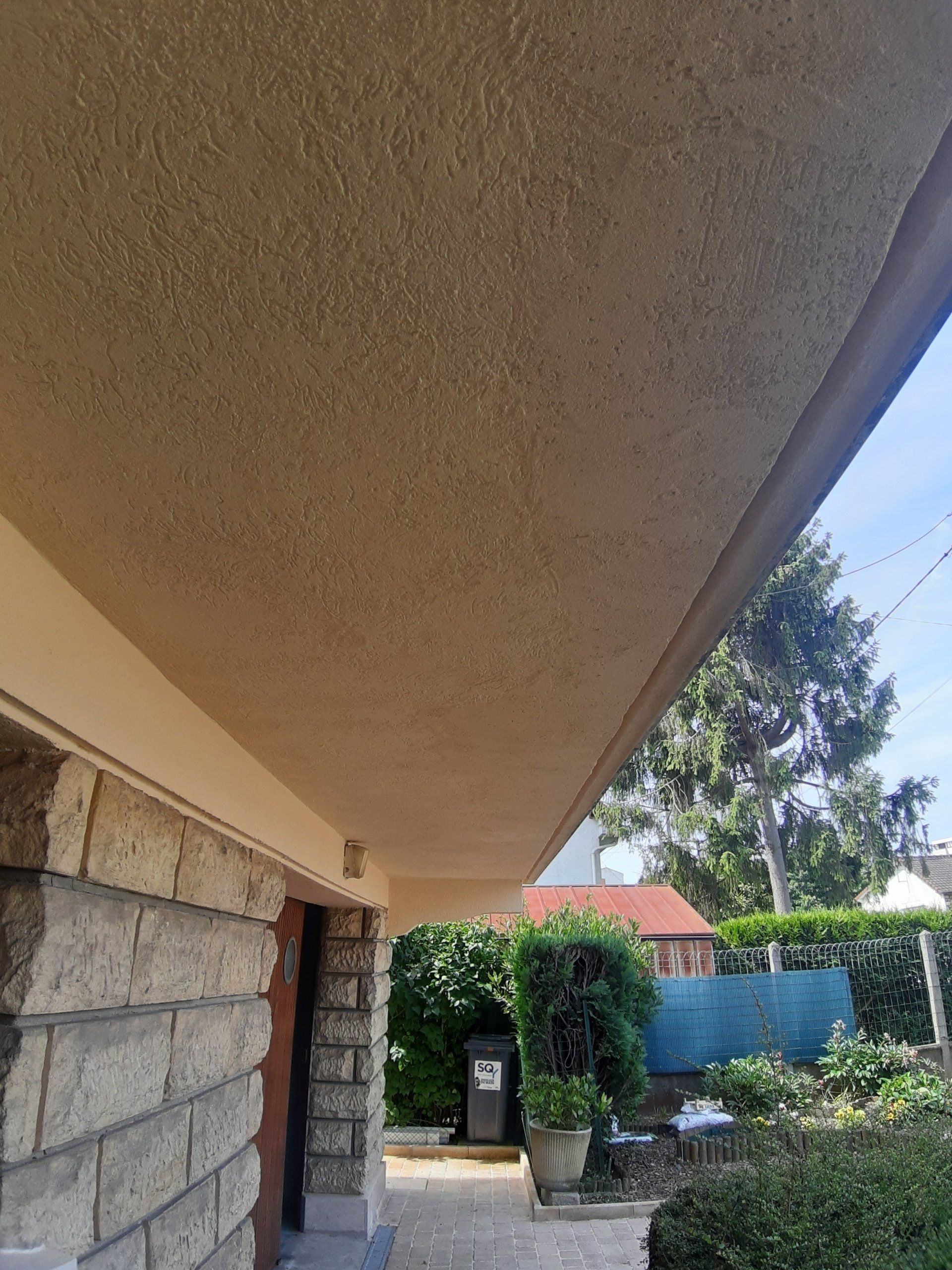 Surplomb texturé beige au-dessus d'une façade en pierre et brique, à côté de verdure et de ciel bleu.