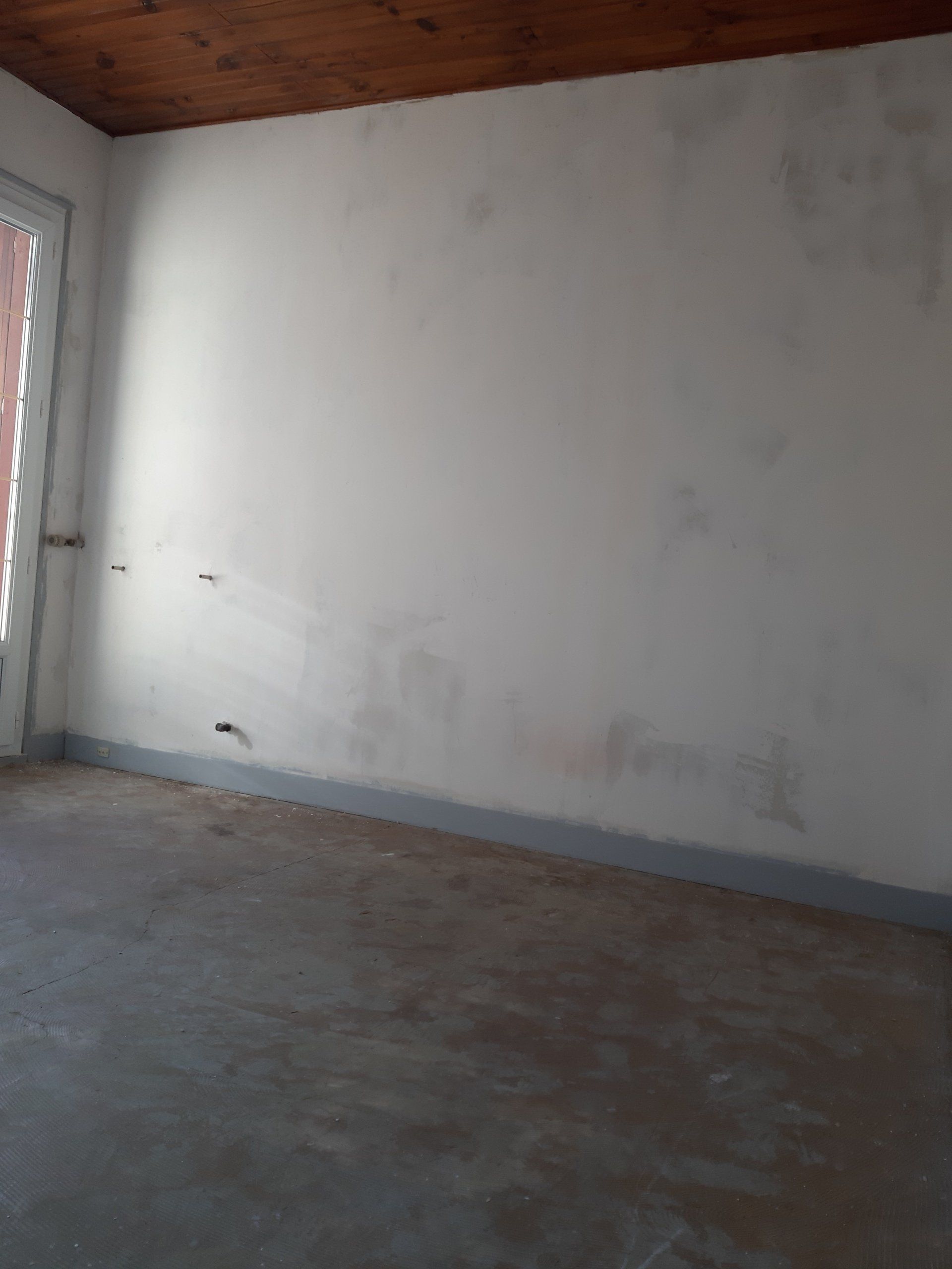 Pièce vide avec des murs blancs, un sol en béton et une porte sur la gauche.