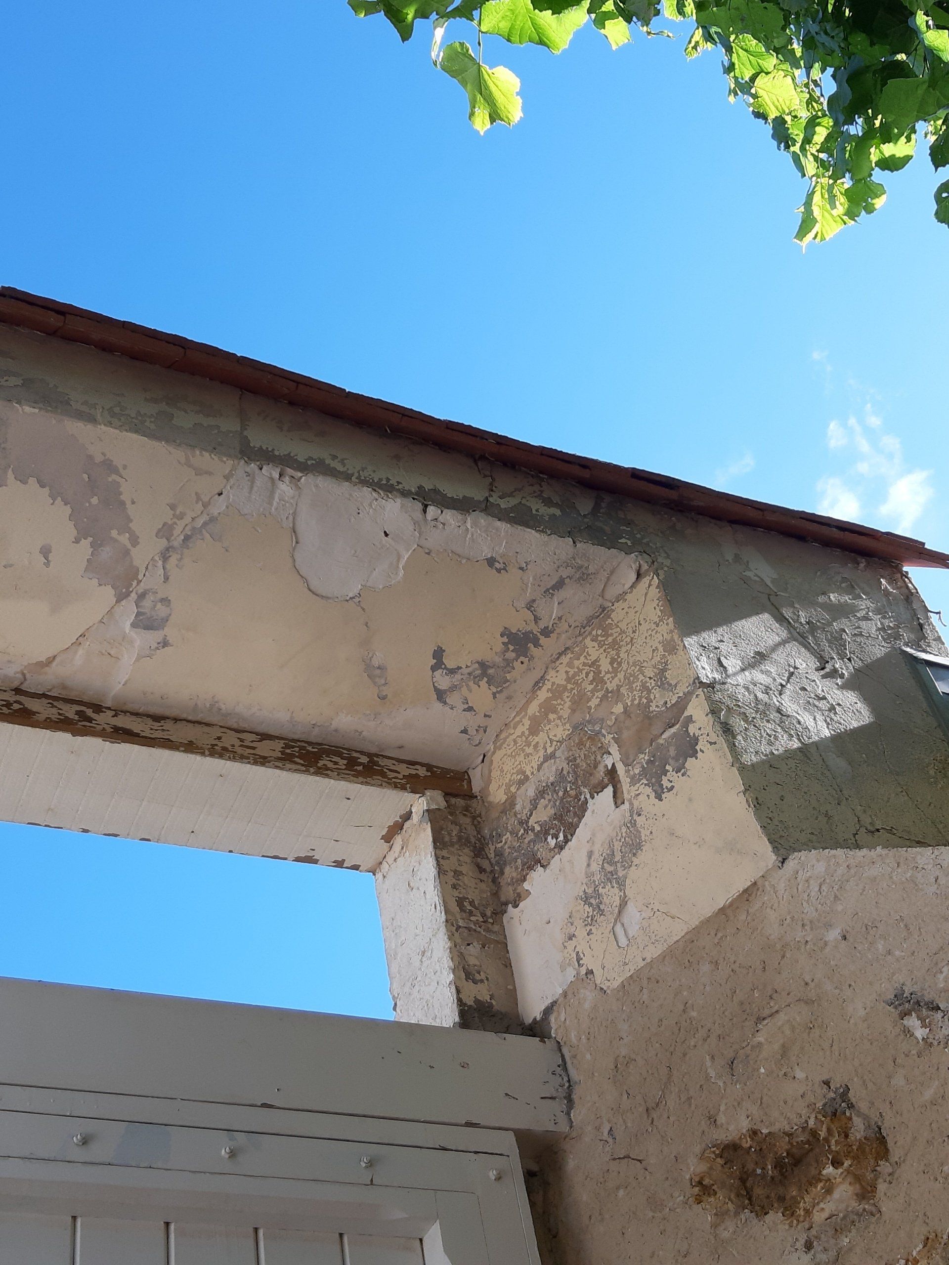 Mur extérieur dont le plâtre s'écaille, révélant les couches sous-jacentes. Un ciel bleu apparaît à travers une fenêtre.