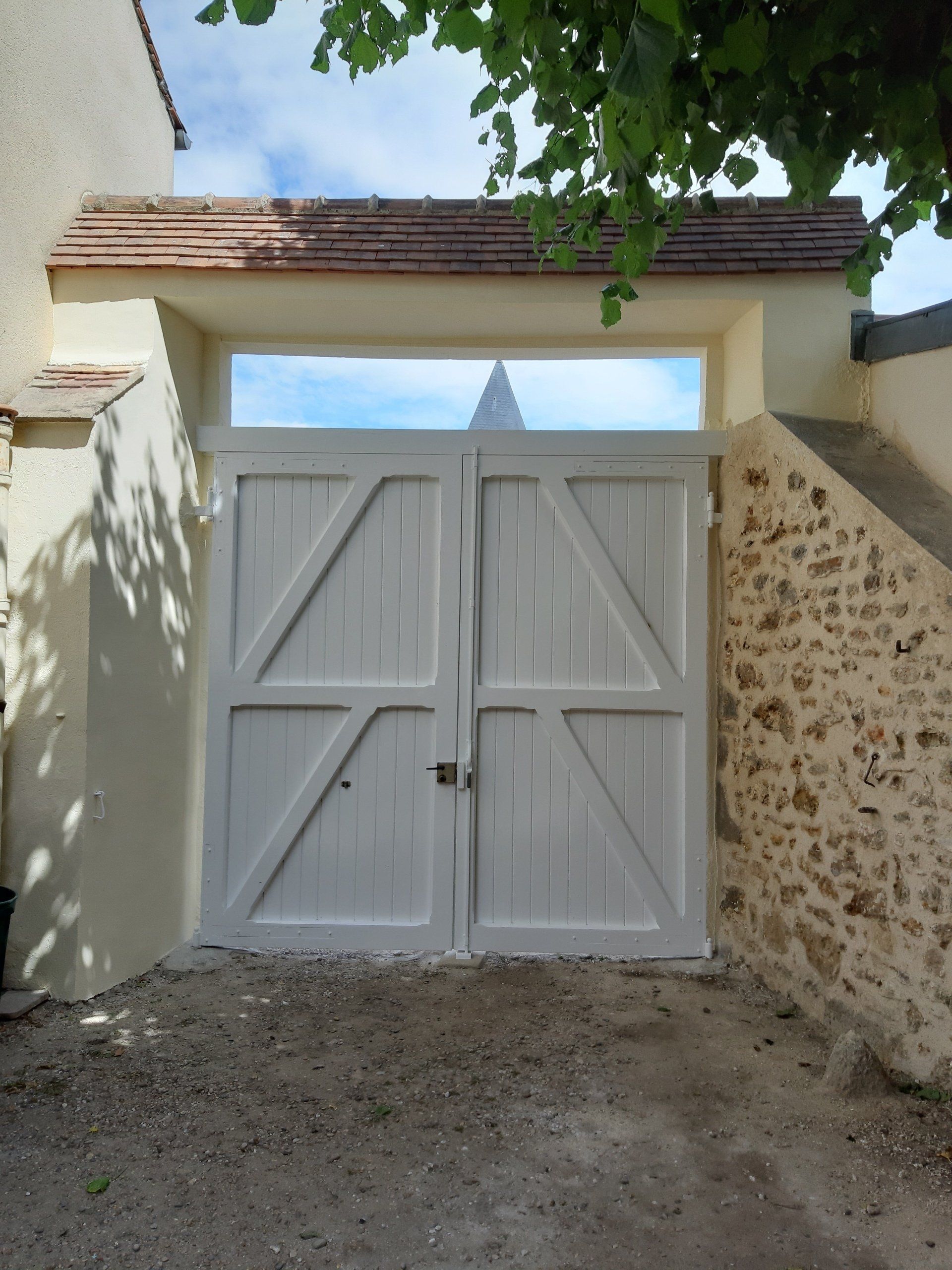 Portail double en bois blanc avec une vue dégagée sur le ciel et un clocher d'église.
