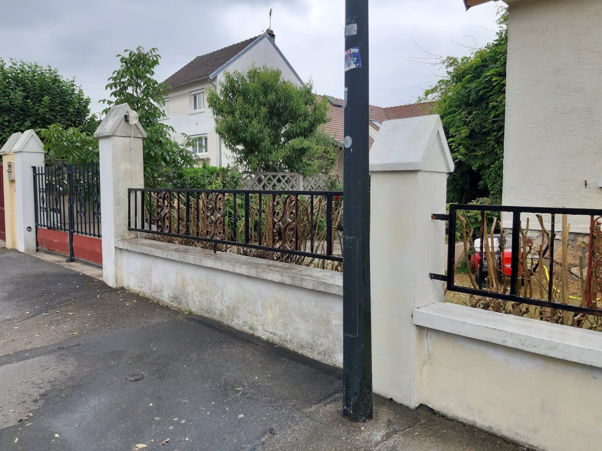 Mur blanc avec clôture métallique noire, portail et lampadaire dans une rue. Arbres verts et bâtiment en arrière-plan.