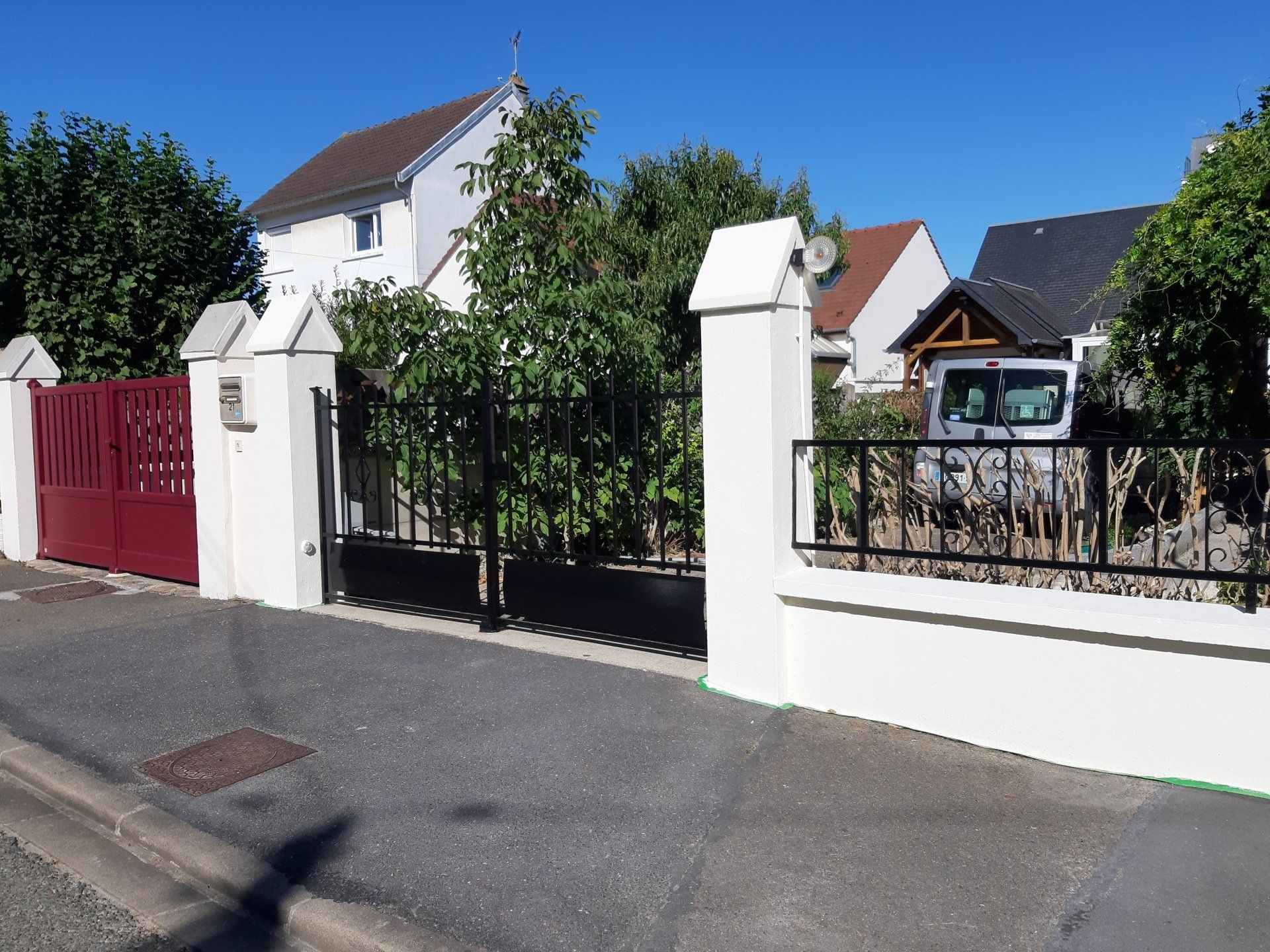 Portail rouge et clôture en fer forgé noir devant une maison blanche avec un ciel bleu.