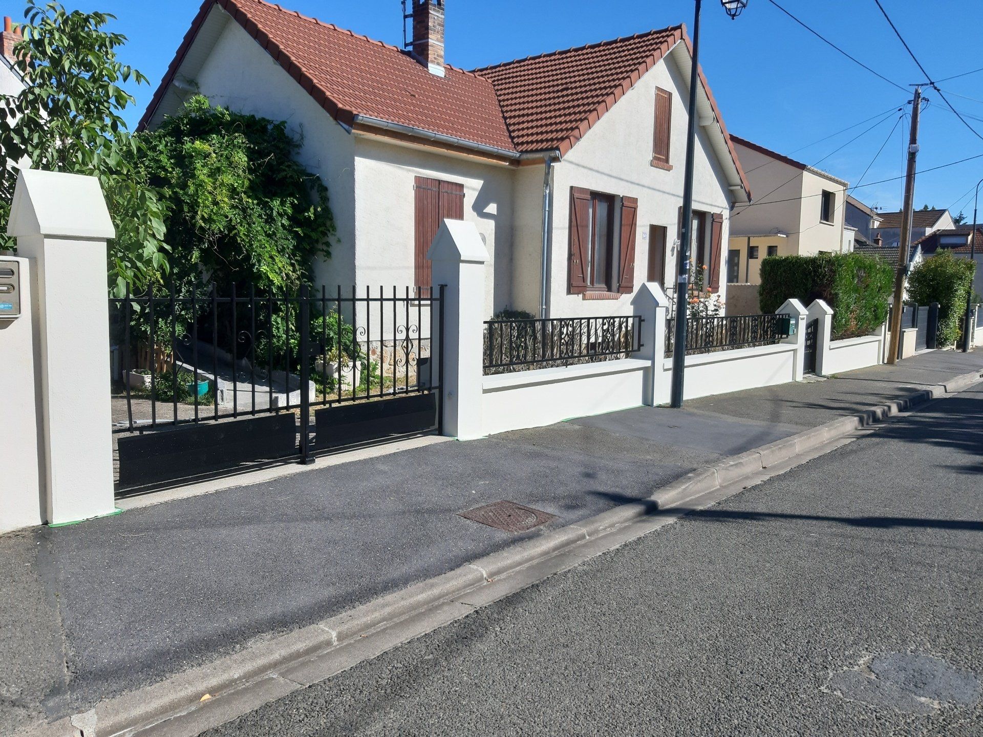 Trottoir devant une maison blanche avec un toit rouge, un portail noir et une clôture environnante par une journée ensoleillée.