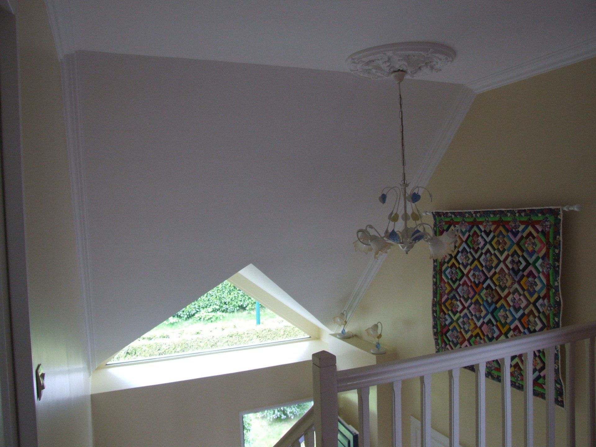 Cage d'escalier avec luminaire, textile suspendu coloré et vue extérieure à travers une fenêtre triangulaire.