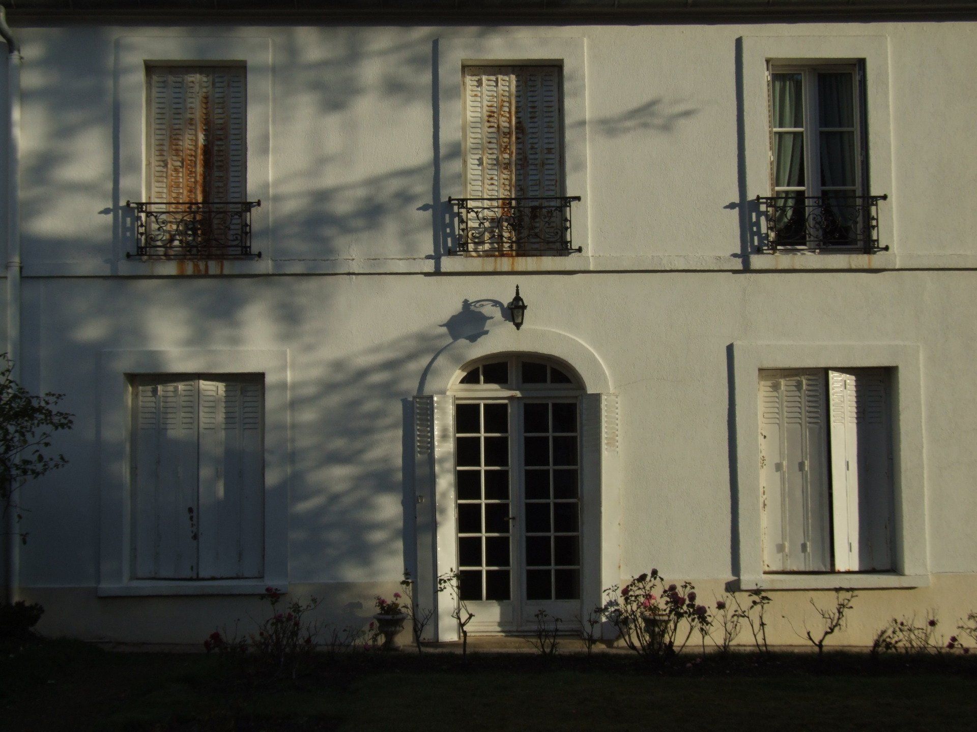 Bâtiment blanc avec porte cintrée et fenêtres avec volets et barreaux de fer rouillés.