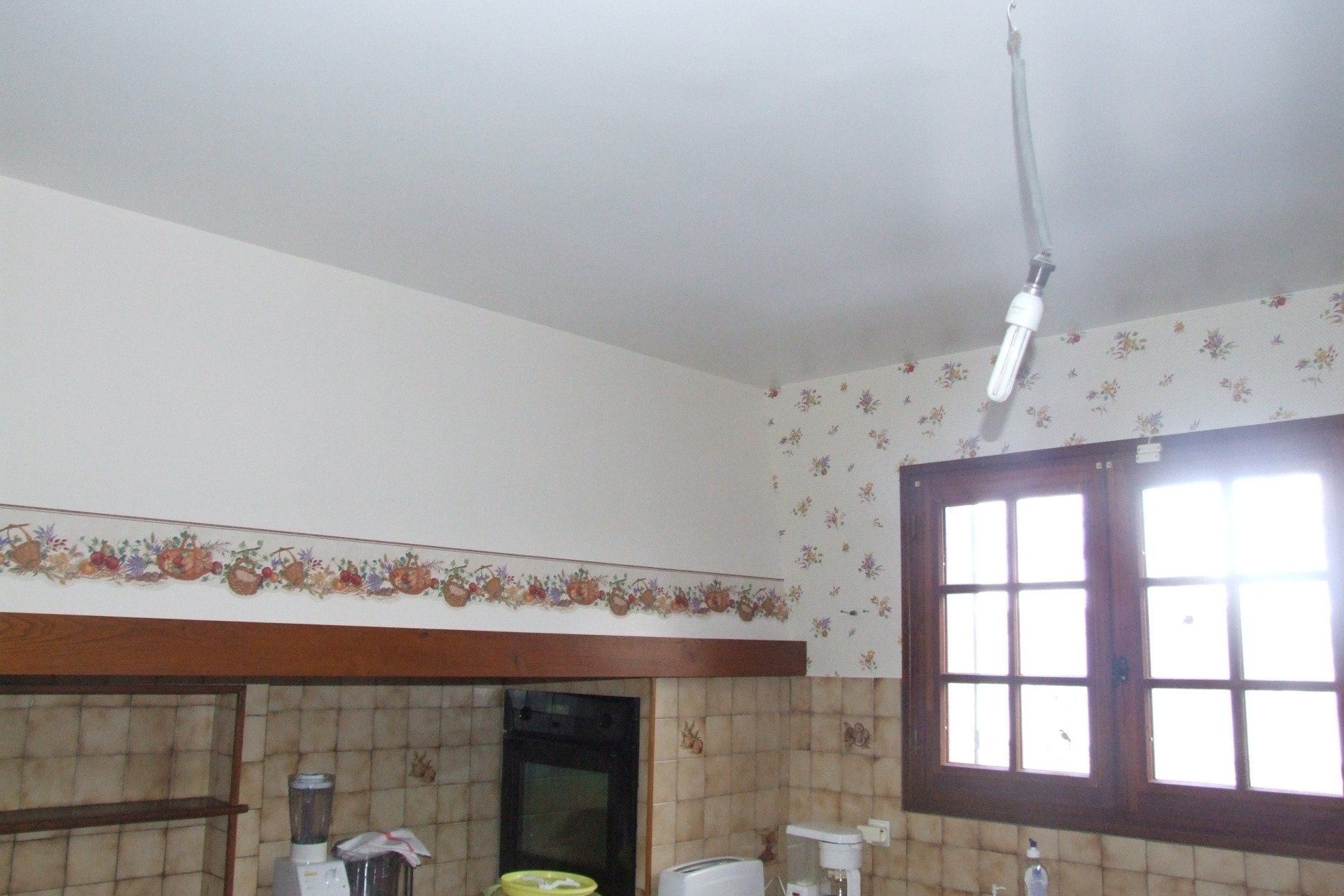 Cuisine avec papier peint fleuri, une fenêtre avec cadre en bois et une suspension lumineuse.