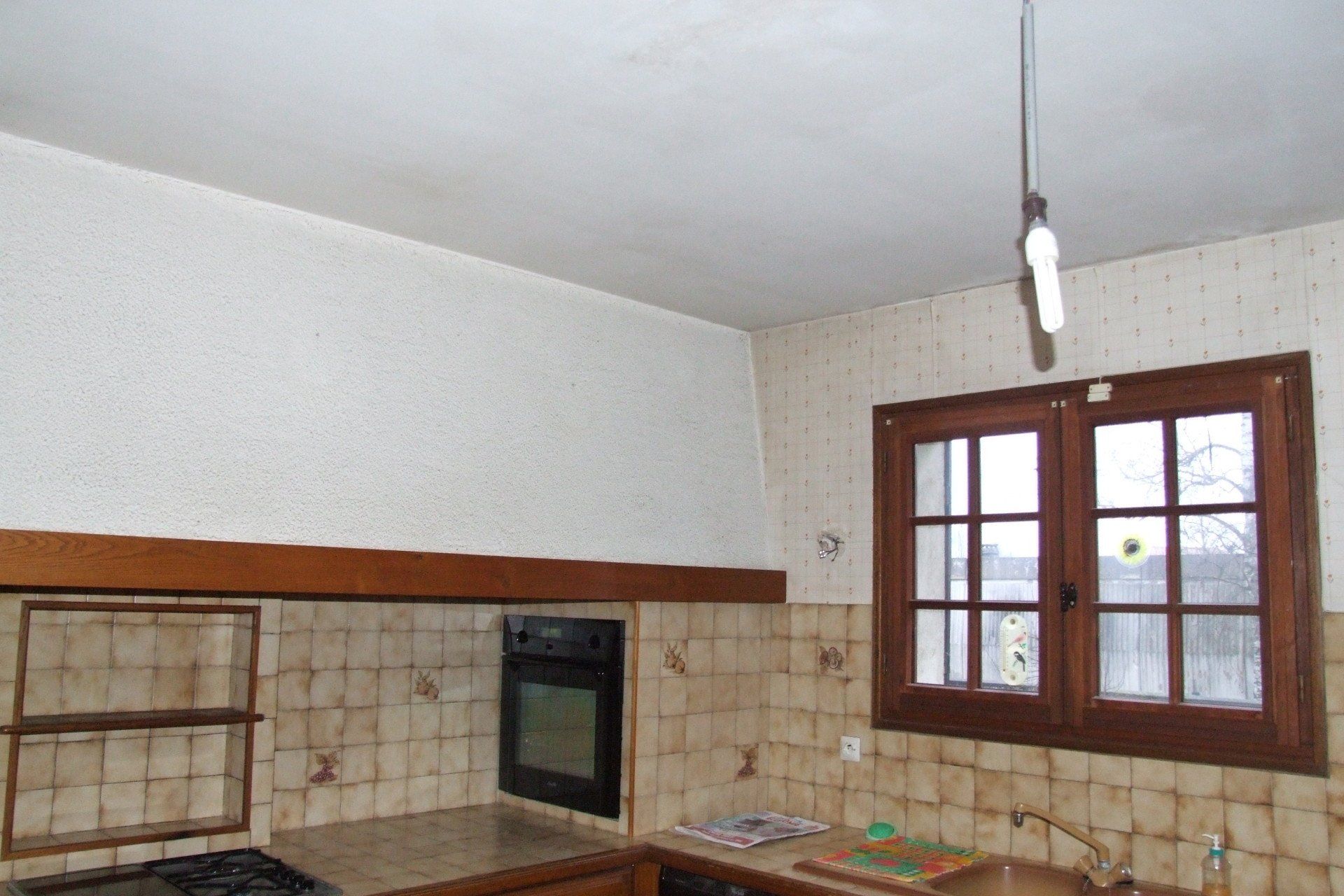 Intérieur de cuisine avec fenêtre, armoires, four et ampoule apparente. Boiseries marron.