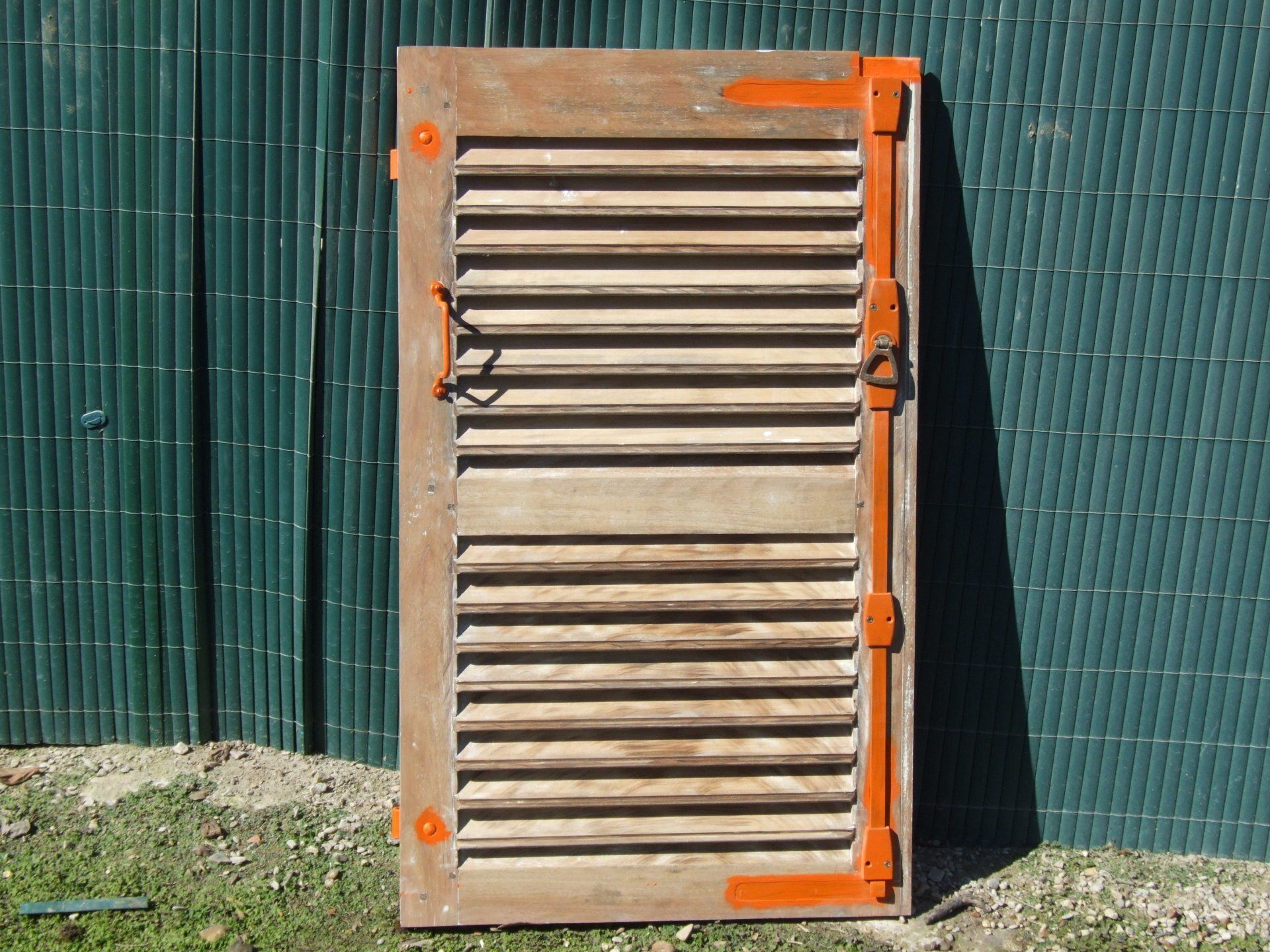 Volet en bois avec charnières et ferrures oranges ; repose contre une clôture verte.