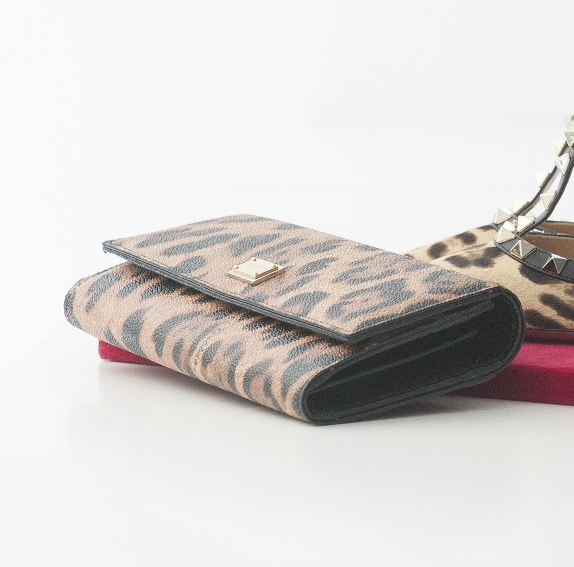 Carteira com estampa de leopardo sobre superfície vermelha, com um sapato ao fundo.