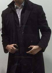 Manequim de sobretudo preto, camisa branca, mãos perto da cintura, em ambiente interno.
