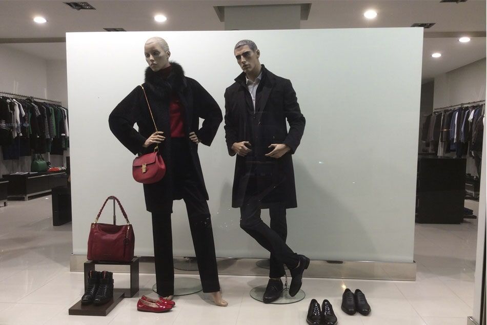 Dois manequins em uma vitrine de loja; mulher de casaco preto, homem de casaco preto, com sapatos e bolsas.