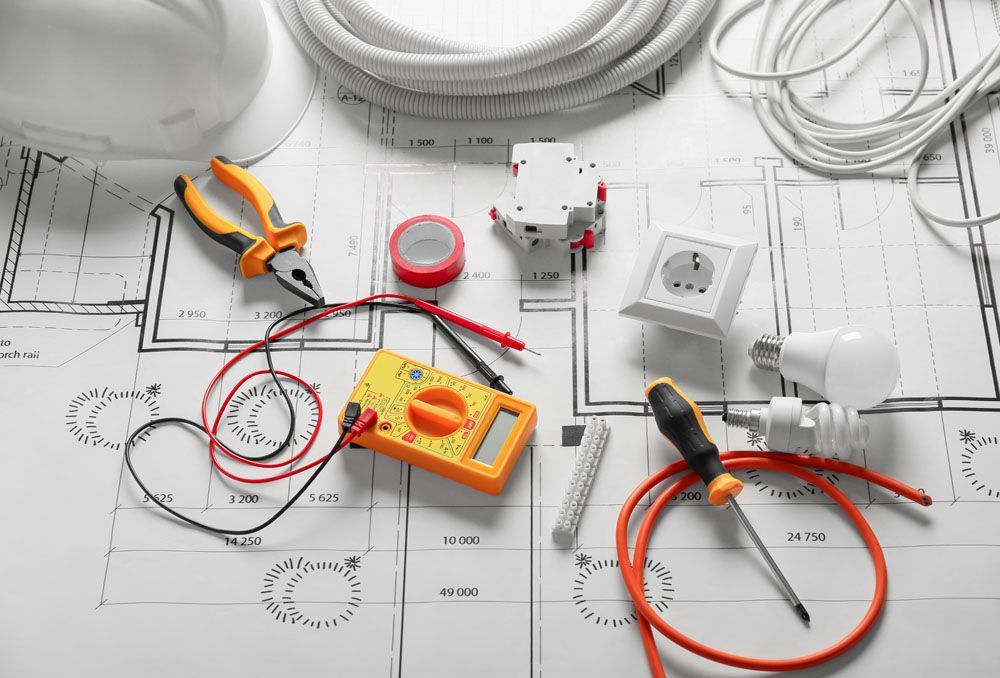 Auf einem Bauplan angeordnete elektrische Werkzeuge und Komponenten, einschließlich Multimeter, Kabel und Steckdose.